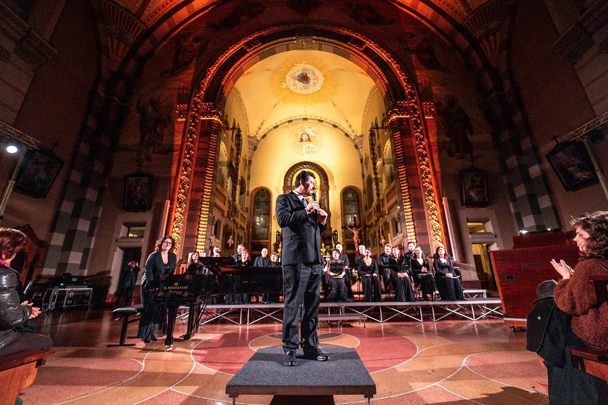 Il M° Gianmario Cavallaro sul podio al centro di una chiesa illuminata di rosso e oro, con il coro e il pianoforte sullo sfondo